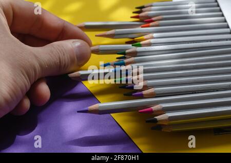Close-up of a man's hand pulling a gray wooden pencil out of a box. Set of multicolored watercolor pencils on a blue-yellow background with shadows. D Stock Photo