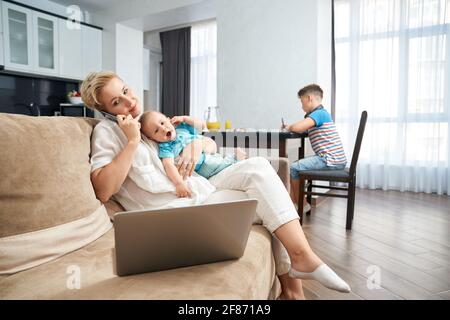 Beautiful woman talking on mobile, working on laptop and holding baby while her eldest son sitting at kitchen table and studying. Concept of motherhood and family time. Stock Photo