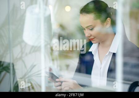 Young smiling businesswoman checking text messages in smartphone when sitting at coffee table, view through window Stock Photo