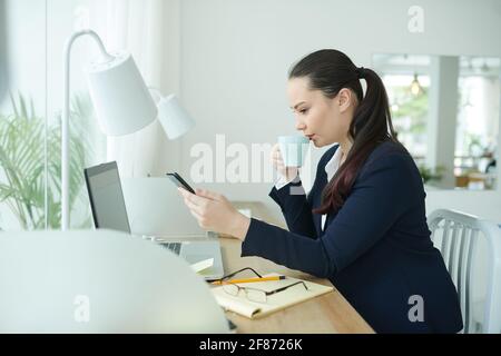 Serious young female entrepreneur drinking coffee and reading text message from coworker or business partner Stock Photo