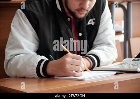 Hands of bearded guy in casualwear bending over desk and making notes or drawing sketch in copybook during lecture in auditorium of university Stock Photo