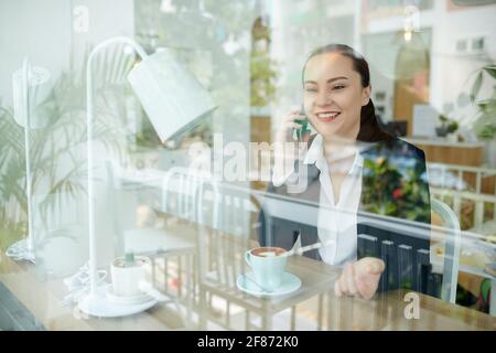 Happy young businesswoman drinking coffee in coffeeshop and talking on phone with friend or colleague, view from outside Stock Photo