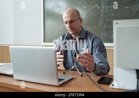 Mature teacher in shirt and eyeglasses sitting by desk, looking at laptop display and talking in microphone during explanation of subject Stock Photo