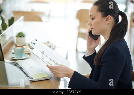 Serious young business lady reading document and talking on phone with colleague Stock Photo