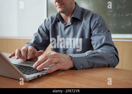 Hands of mature man in grey shirt touching buttons of laptop keypad while sitting by desk against blackboard and entering data Stock Photo