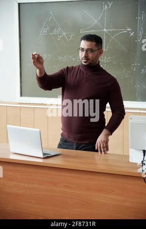 Mixed-race male in casualwear holding piece of chalk while standing between desk and blackboard and explaining something to students Stock Photo