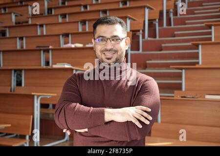 Young successful male teacher of university crossing his arms by chest while standing in front of camera against desks in lecture hall Stock Photo