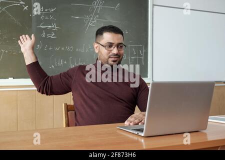 Mixed-race male teacher in casualwear pointing at solution of equation on blackboard while sitting by desk and looking at laptop display Stock Photo