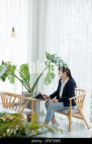 Young businesswoman sitting at cafe table, drinking coffee and using application on tablet computer Stock Photo
