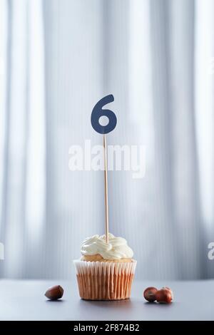 Anniversary or birthday digital greeting card concept. Tasty vanilla cupcake or muffin with number 6 six on white plate with hazelnuts nearby with bright background. High quality vertical photo Stock Photo