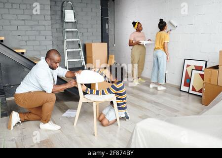 Young father and little son assembling wooden chair on the floor against mother and daughter painting wall of living-room in white color Stock Photo