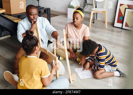 Puzzled African man looking at parts of wooden chair while going to assemble it, woman and boy pointing at assembling instruction Stock Photo