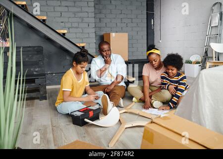 Puzzled African man looking at parts of wooden chair on the floor while his wife and little son looking through assembling instruction Stock Photo