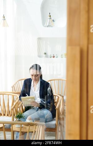 Creative young businesswoman sitting in armchair in coffeeshop and taking notes in planner Stock Photo