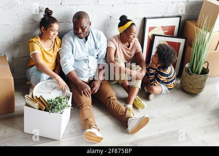 Young cheerful family of father, mother, little son and teenage daughter talking while sitting against wall on the floor of living-room Stock Photo
