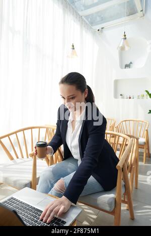 Positive young pretty woman running online business, checking feedback from clients and drinking coffee in coffeeshop Stock Photo