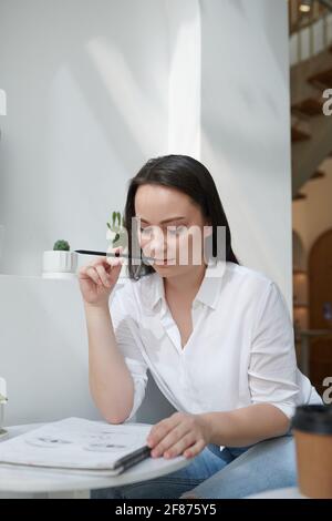 Pensive pretty creative young woman biting pencil and looking at portrait she is drawing fro friend Stock Photo
