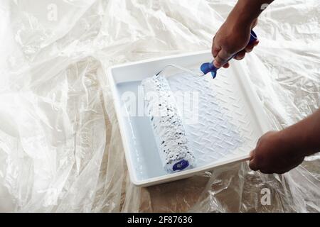 Hands of young African man holding paint-roller while taking some paint from white square plastic container during home renovation Stock Photo
