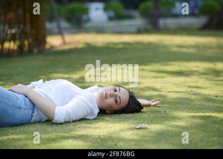 Attractive young brunette relaxing on green grass in city park and smiing at camera Stock Photo