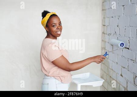 Happy young African female with paintroller standing in the corner in front of brick wall while painting it into white color during home renovation Stock Photo