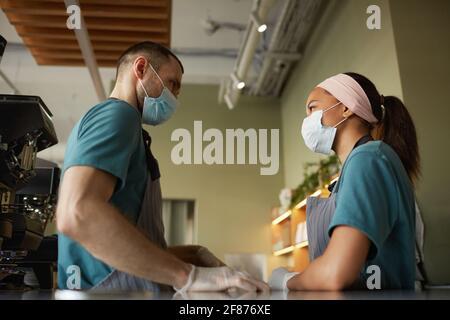 Side view portrait of two young workers wearing masks while standing at bar counter in cafe or coffee shop Stock Photo