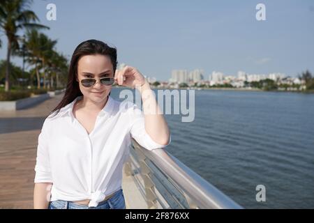 Portrait of young beautiful woman taking off sunglasses and smiling at camera when standing on river bank Stock Photo