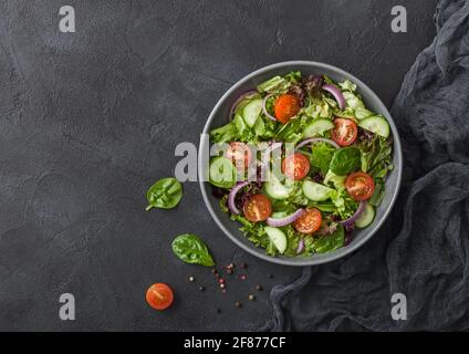 Fresh healthy vegetarian vegetables salad with tomatoes and cucumber, red onion and spinach in grey bowl plate on dark background with black kitchen c Stock Photo