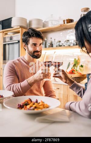 Cute multi-ethnic couple having lunch together in the kitchen and drinking glass of wine. Stock Photo