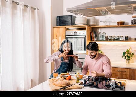 Cheerful black woman and her boyfriend having delicious breakfast in the kitchen at home and taking photo. Stock Photo