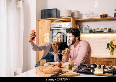 Cheerful black woman and her boyfriend having delicious breakfast in the kitchen at home and taking selfie. Stock Photo