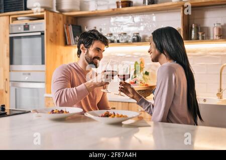 Cute multi-ethnic couple having lunch together in the kitchen and drinking glass of wine. Stock Photo