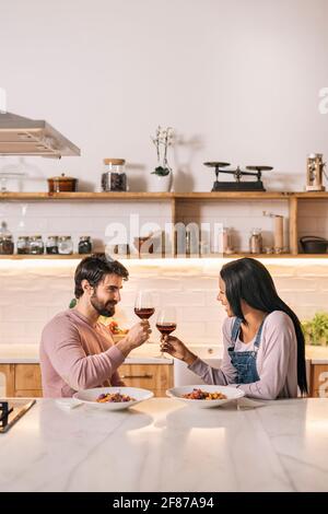 Cute multi-ethnic couple having lunch together in the kitchen and drinking glass of wine. Stock Photo