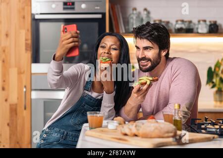 Cheerful black woman and her boyfriend having delicious breakfast in the kitchen at home and taking selfie. Stock Photo