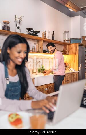 Beautiful black woman using her laptop in the kitchen while her boyfriend cooks. Stock Photo
