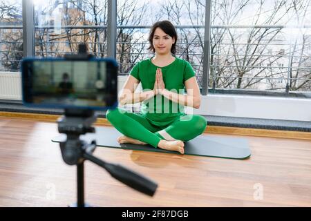 Female fitness trainer in green sportswear sit at mat in lotus pose with namaste gesture to start remote yoga exercise workout training Stock Photo