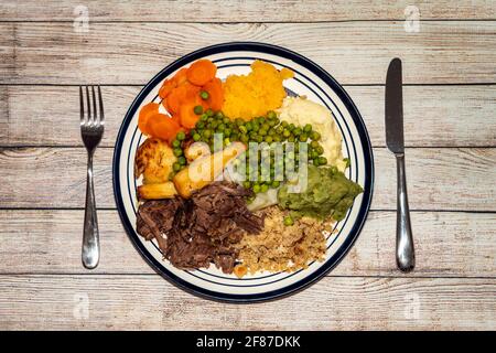 Sunday dinner of slow cooked shredded pulled beef on a plate with a knife and fork and plenty of vegetables such as peas, mushy peas, parsnips, roast Stock Photo