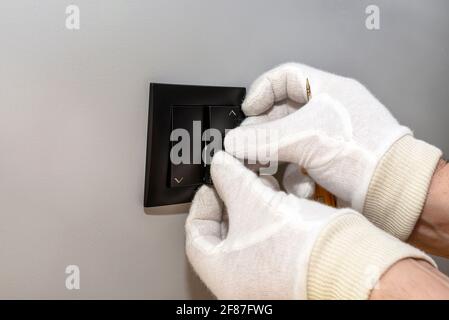 An electrician mounts the black button of the roller shutter switch up and down with white gloves in the room by the window. Stock Photo