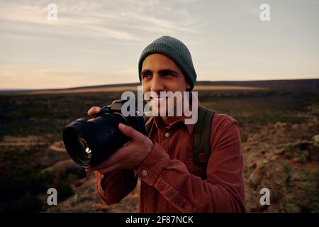Young photographer with backpack and cap holding professional camera and shooting morning sunrise Stock Photo