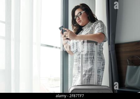 Serious young woman standing in hotel room next to her luggage and using mobile application on smartphone to order transfer or taxi to airport Stock Photo