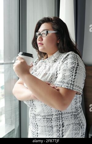 Pensive young woman in glasses drinking morning decaf coffee and looking through window of hotel room Stock Photo