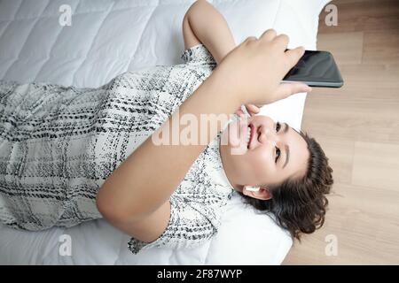 Lovely pretty young woman wearing earbuds when lying on bed and video calling her friend or family member Stock Photo