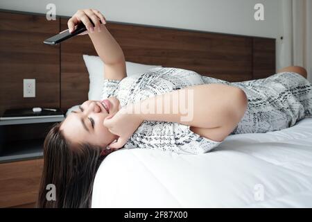 Smiling pretty young woman lying on bed in hotel room and video calling her friend or boyfriend Stock Photo