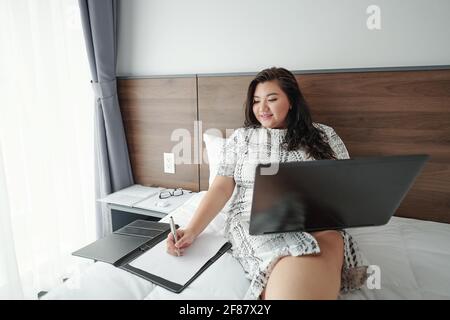 Smiling pretty young businesswoman sitting on bed , working on laptop and taking notes in document Stock Photo