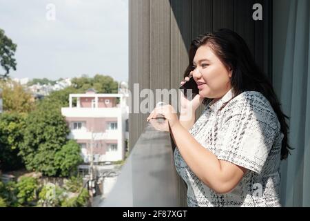 Pretty young plus size woman standing on balcony and talking on phone with friend or family member Stock Photo