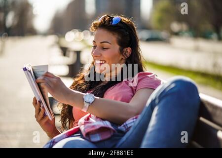 Happy young woman is enjoying sunny day outdoor in city. She is reading book. Stock Photo