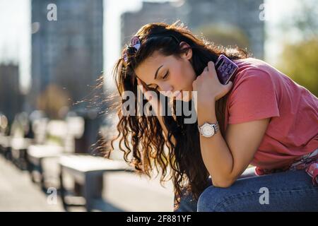 Sad young woman is sitting on bench in city. She is depressed.