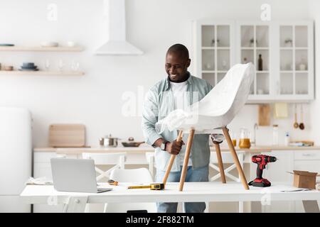 Order online. Smiling black man receiving new chair, checking it and looking at laptop in kitchen interior Stock Photo