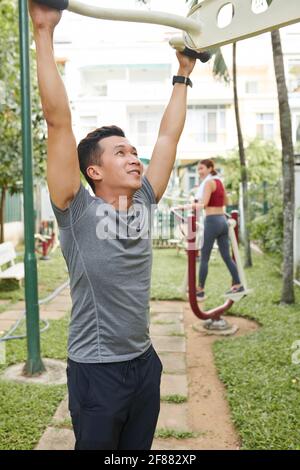 Handsome fit young Asian man doing exercises on fitness equipment in city park Stock Photo