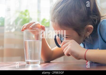 The child examines the water with a magnifying glass in a glass. Selective focus. Kid. Stock Photo