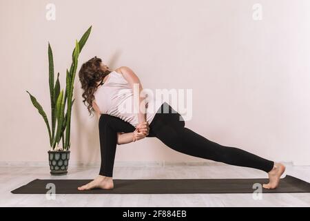 A woman in sportswear practicing yoga, doing parivritta parshvakonasana, reverse angle pose, practicing in the studio against the wall on a mat. Stock Photo